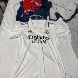 Vit Real Madrid fotbollströja - Snygg vit Real Madrid fotbollströja från Adidas med korta ärmar. Tröjan har klubbens emblem och sponsorlogga på framsidan. Perfekt för alla fotbollsfans som vill visa sitt stöd för laget. Äkta kostar 1400 ny kan diskurera pris