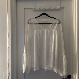 Vit blus från Vila - Säljer en elegant vit blus från Vila med v-ringning och spetsdetaljer vid axlarna. Blusen har långa ärmar och ett lätt transparent material med diskret prickigt mönster. Perfekt för en stilren look.