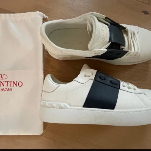 Valentino Garavani sneakers i vitt och svart - Snygga Valentino Garavani sneakers i vitt med en bred svart rem över ovansidan. Skorna har en stilren design med en tjock sula och subtila detaljer. Perfekta för en trendig look. Givetvis äkta och påse finns 