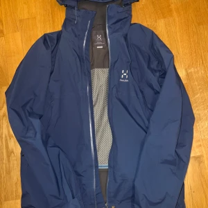 Blå vindjacka från Haglöfs - Säljer en blå vindjacka från Haglöfs med dragkedja och huva. Perfekt för utomhusaktiviteter med sin funktionella design och slitstarka material. Jackan har en stilren look och är idealisk för både vår och höst.