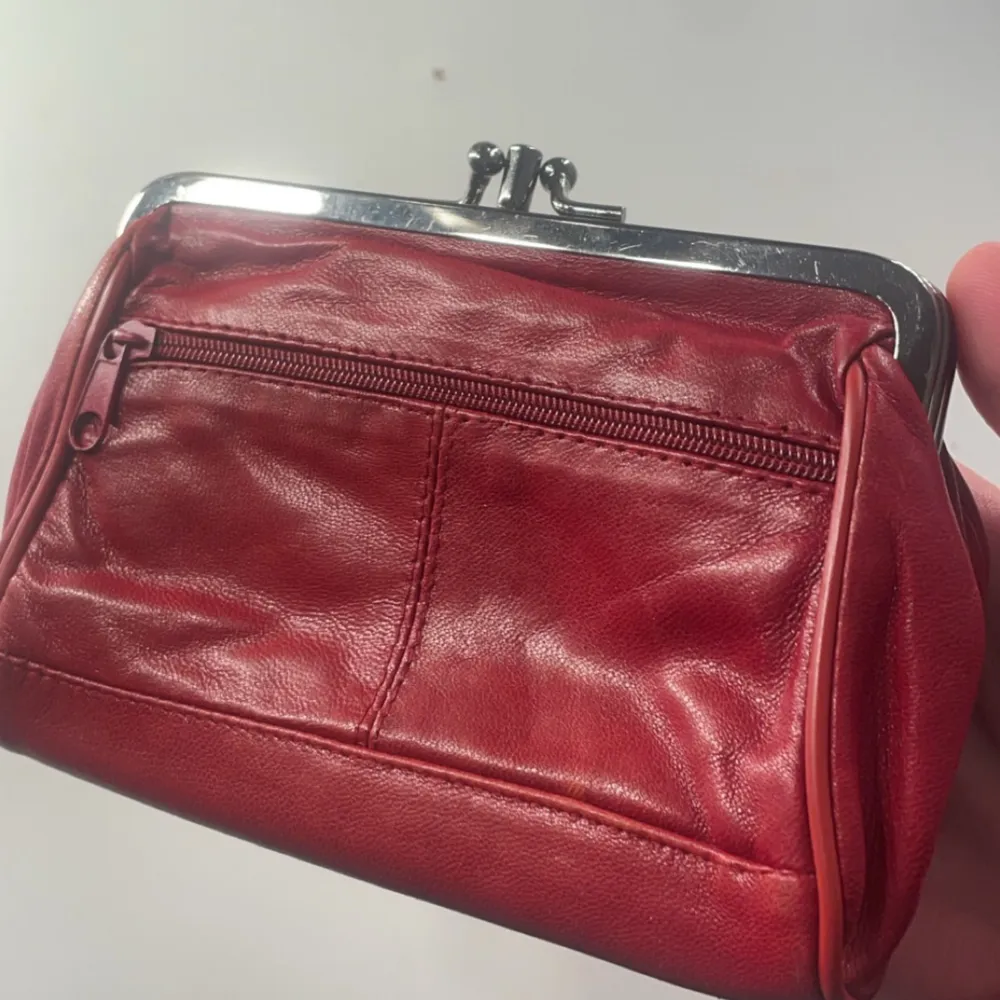 Snygg röd plånbok i skinn med en praktisk dragkedja på framsidan. Den har en klassisk metallram och ett rymligt fack inuti. Perfekt för att hålla ordning på dina kort och kontanter. Riktig läder. Laukut & Käsilaukut.