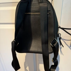 Svart ryggsäck med dragkedja - Snygg svart ryggsäck i skinn med guldfärgade dragkedjor. Den har justerbara axelremmar och en praktisk ytterficka med dragkedja. Perfekt för att bära dina dagliga nödvändigheter med stil.