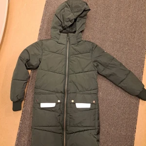 Grön lång dunjacka från Everest - Säljer en grön lång dunjacka från Everest med huva och dragkedja. Jackan har praktiska fickor framtill och är perfekt för kalla dagar. Den är quiltad och har långa ärmar med muddar.