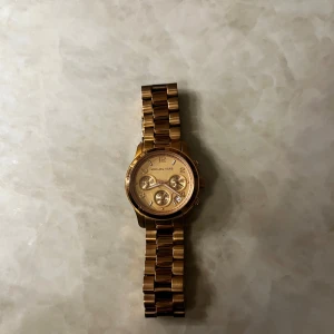 Guldfärgad klocka från Michael Kors - Snygg guldfärgad klocka från Michael Kors med metallarmband och flera urtavlor. Perfekt accessoar för att lyfta din stil. Klassisk design med en modern touch.