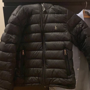 Svart dunjacka från Polo Ralph Lauren - Snygg svart dunjacka från Polo Ralph Lauren med dragkedja och två fickor framtill. Jackan har en klassisk design med långärmade ärmar och är perfekt för kyligare väder. Kan sänka pris vid snabb affär