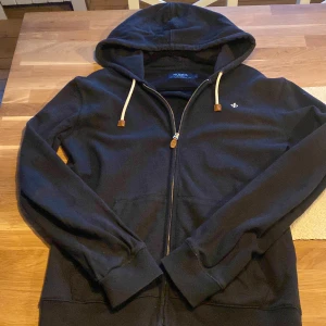 Marinblå zip hoodie från Morris - Säljer en stilren marinblå zip från Morris med dragkedja och en liten broderad detalj på bröstet. Tröjan har långa ärmar och en justerbar huva med vita snören. Perfekt för en avslappnad look. Om det är något du undra så är det bara att skriva till mig 