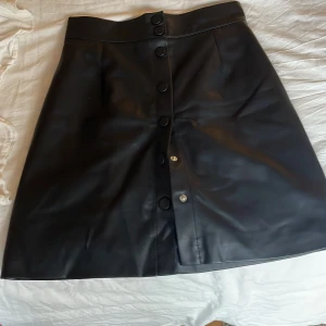 Svart läder kjol med knappar - Snygg svart kjol i skinnimitation med knappar framtill. Kjolen har en stilren design och är perfekt för att ge en edgy touch till din outfit. Den är i en klassisk kort modell och passar bra till olika tillfällen.