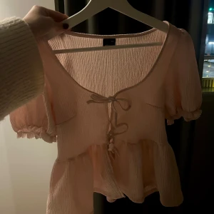 Rosa blus med knyt och volangdetaljer - Söt rosa blus med korta puffärmar och knyt framtill. Blusen har volangdetaljer och en lätt ribbad struktur, perfekt för vår och sommar💗💗💗