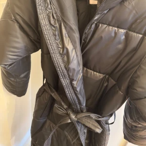 Svart dunjacka från Nelly - Säljer en svart dunjacka från Nelly med bälte i midjan. Jackan har en lång design och storlek xxs helt oanvänd