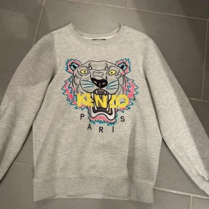 Grå sweatshirt från Kenzo - Säljer en grå sweatshirt från Kenzo med ett stort, färgglatt tigertryck på framsidan. Tröjan har långa ärmar och rund halsringning. Perfekt för en avslappnad stil med en touch av lyx.
