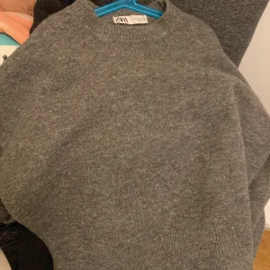 Grå stickad tröja från Zara - Säljer en mysig grå stickad tröja från Zara. Perfekt för kyliga dagar med sin klassiska design och ribbade detaljer vid ärmslut och nederkant. Enkelt att matcha med olika outfits.