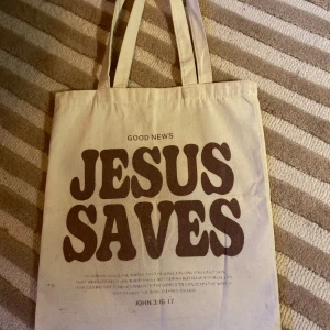 Tygväska med text   - Säljer en tygväska med texten JESUS SAVES' tryckt i stor stil. Väskan har långa handtag och är perfekt för att bära med sig det nödvändigaste. Tillverkad i ett slitstarkt material som passar för daglig användning. Normal storlek 