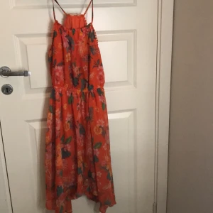 Orange blommig klänning från H&M - Säljer en färgglad orange klänning från H&M med blommigt mönster i rosa, orange och grönt. Klänningen har smala axelband och en luftig passform, perfekt för varma dagar. Den har fina detaljer med volanger längs kanterna. Har några små fläckar längs baksidan, uppe vid nacken som syns på sista bilden.