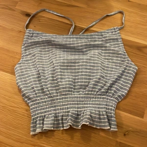 Randig topp från Gina Tricot - Säljer en söt randig topp från Gina Tricot i blått och vitt. Toppen har smala axelband och en elastisk midja med volangdetalj. 