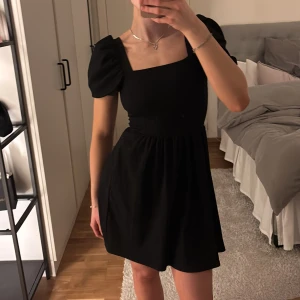 Svart klänning med puffärmar - Snygg svart klänning med puffiga kortärmar och fyrkantig urringning. Perfekt för en stilren look. Klänningen har en åtsittande midja och en lätt utsvängd kjol som ger en fin silhuett.