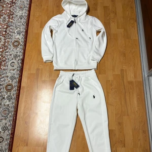 Vit hoodie och byxor från Ralph Lauren - Säljer en stilren vit hoodie och matchande byxor från Ralph Lauren. Hoodien har dragkedja och ribbade muddar, medan byxorna har en elastisk midja. Båda plaggen pryds av den ikoniska logotypen. Perfekt för en avslappnad look.