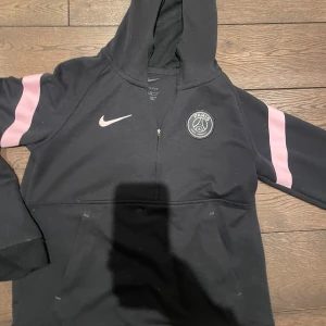 Svart hoodie från Nike med PSG-logga - Snygg svart hoodie från Nike med Paris Saint-Germain-logga på bröstet. Den har rosa ränder på ärmarna och en praktisk dragkedja framtill. Skriv vid frågor och bilder, pris kan diskuteras vid snabb affär då jag behöver pengar