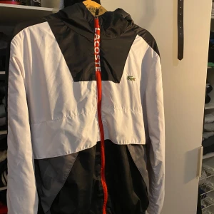 Svart och vit vindjacka från Lacoste - Snygg svart och vit vindjacka från Lacoste med röd dragkedja och logga på bröstet. Jackan har en huva och stora bokstäver på ryggen. Perfekt för en sportig look.