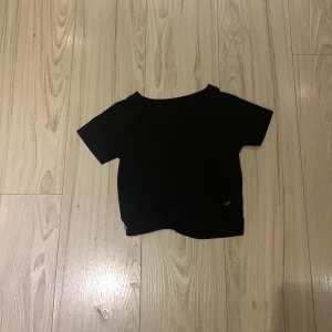 Svart kortärmad topp med knutdetalj - Snygg svart kortärmad topp med en knutdetalj framtill. Perfekt för en stilren look. Liten logga i nederkant.