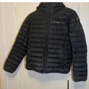 Svart dunjacka från Peak Performance - Snygg svart dunjacka från Peak Performance med dragkedja och huva. Perfekt för kyliga dagar med sin quiltade design som håller värmen. Jackan har en stilren look och är lätt att matcha med olika outfits.
