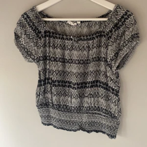 Mönstrad topp i svart och vit - Säljer en snygg mönstrad topp i svart och vit med korta ärmar. Toppen har ett elastiskt band vid halsen och nederkanten för en bekväm passform. Perfekt för en avslappnad stil.