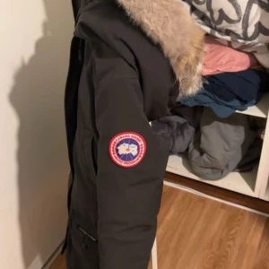 Svart parkajacka från Canada Goose - Säljer en svart parkajacka från Canada Goose med en mysig pälsfodrad huva. Jackan har en klassisk design med dragkedja och knappar framtill samt en broderad logotyp på ärmen. Köpte den för 13 tusen förra sommaren.
