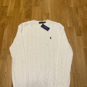 Vit stickad tröja från Polo Ralph Lauren - Säljer en stilren vit stickad tröja från Polo Ralph Lauren med kabelstickat mönster och den klassiska loggan broderad i blått på bröstet. Tröjan har långa ärmar och rund halsringning. Perfekt för en elegant och avslappnad look.