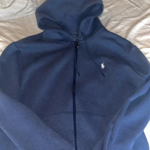 Blå Ralphlauren zip hoodie - Säljer en snygg  hoodie från Polo Ralph Lauren med dragkedja och den klassiska logotypen. Finns inget fel på hoodien varken märken eller slitage. Köpt för 2300 och säljer nu för 1200 pris går att diskutera.