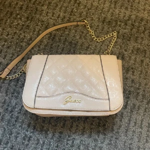 Ljusrosa Guess väska - Snygg ljusrosa  axelväska från Guess med quiltad design och guldfärgad kedja. Väskan har en elegant logotyp framtill och är perfekt för att lyfta din stil. Den är både chic och praktisk med en justerbar rem.