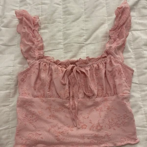 Rosa blommig topp från Shein - Söt rosa topp från Shein med blommigt mönster och volangdetaljer på axelbanden. Toppen har en knytning framtill och är perfekt för en somrig look. Den är tillverkad i ett lätt och luftigt material.