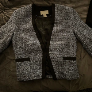 Grå kavaj från H&M - Snygg grå kavaj från H&M med svart kantdetalj. Den har en klassisk design med långärmad stil och två fickor framtill. Perfekt för en stilren look.