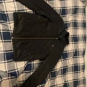 Svart quiltad jacka från Gant - Snygg blå quiltad jacka från Gant med dragkedja och rutig insida. Perfekt för höst och vår. Jackan har långa ärmar och en stilren design med Gant-logga på bröstet. Storlek S använt fåtals gånger 