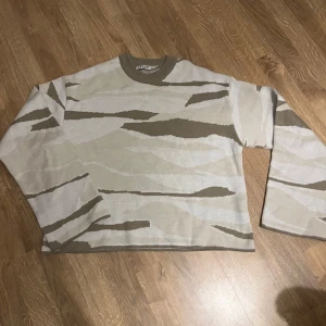 Beige och vit tröja från NA-KD - Säljer en snygg beige och vit tröja från NA-KD med ett coolt kamouflagemönster. Tröjan har långa ärmar och en rund halsringning. Perfekt för en avslappnad stil.