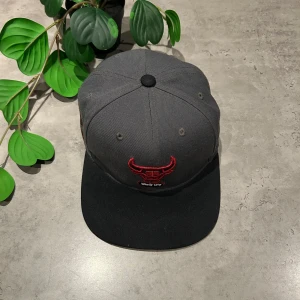 Grå keps med röd broderad logga - Snygg grå keps med svart skärm och röd broderad logga framtill. Kepsen har en klassisk design med justerbar passform och är perfekt för en casual look.
