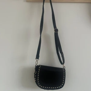 Svart axelväska med nitar - Snygg svart axelväska med coola nitar runt kanterna. Väskan har en justerbar axelrem och är perfekt för att ge din outfit en edgy touch. Tillverkad i syntetmaterial.