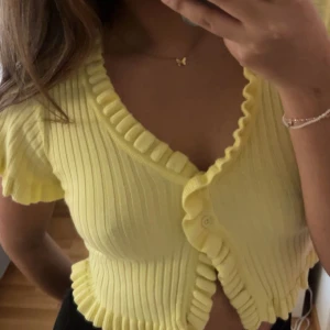 Gul ribbad volang topp från Chiquelle - Säljer en söt gul ribbad topp från Chiquelle med volangdetaljer och knappar framtill. Toppen har korta ärmar och en croppad passform. Perfekt att ha själv eller över en klänning - som en kofta. Säljer för att den inte kommer till användning.