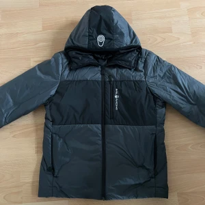 Svart och grå pufferjacka från Sail Racing - Snygg svart och grå pufferjacka från Sail Racing med huva och dragkedja. Jackan är vindtät och andas, perfekt för kyliga dagar. Den har en stilren design med logotypdetaljer på bröstet och huvan. Jackan har inga skador och har använts en vinter. Den är storlek M men passar mer på folk med storlek S och XS