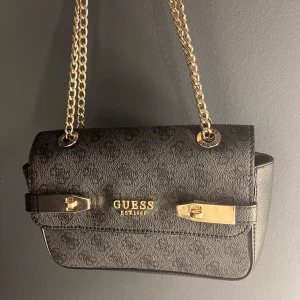 Guess väska - Säljer min så snygga svarta axelväska från Guess med guldfärgade kedjehandtag och detaljer. Väskan är rymlig och har praktiska fack för förvaring. Köptes på zalando och är använd fåtal gånger men i bra skick❤️Pris kan diskuteras vid snabb affär!
