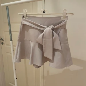 Beige kjol med knyt från Zara - Snygg beige kortkjol från Zara med volangdetaljer och ett knytband i midjan. Perfekt för en stilren look.