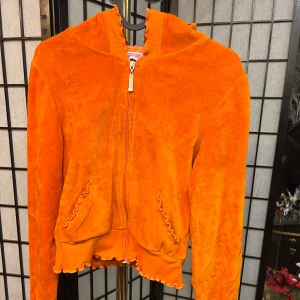 Unik orange cropped hoodie från Tone Shore - Säljer en unik orange hoodie från Tone Shore med dragkedja och volangdetaljer. Perfekt för en avslappnad stil med en touch av färg. Den har långa ärmar och en mysig huva.