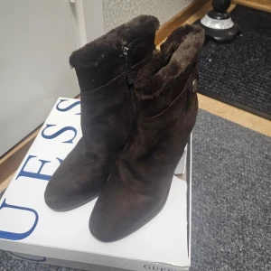 Bruna boots med kilklack från Guess - Snygga bruna boots från Guess med kilklack och mjukt foder. Skorna har en dragkedja på sidan och en dekorativ knappdetalj. Perfekta för kyligare dagar.