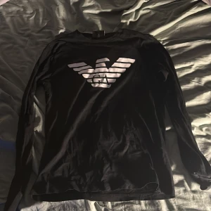 Svart långärmad tröja från Emporio Armani - Snygg svart långärmad tröja från Emporio Armani med det ikoniska logotyptrycket på framsidan. Perfekt för en stilren look.