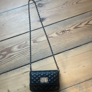 Svart axelväska med kedja från glitter som är använd 1 gång - svart axelväska med en  kedjerem. Väskan har en praktisk dragkedja inuti och ett metallspänne framtill. 