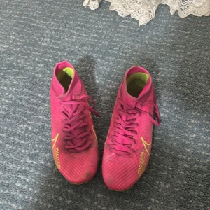 Rosa Nike fotbollsskor - Säljer ett par rosa Nike fotbollsskor med snörning och gula detaljer. Skorna har en hög krage för extra stöd och är designade för optimal prestanda på planen. Storlek 42