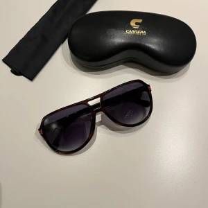 Solglasögon från Carrera - Snygga solglasögon från Carrera med en klassisk pilotform. Bågarna är i en mörkbrun nyans med svarta detaljer och glasen är tonade. Perfekt för en stilren look. Levereras med originalfodral och putsduk. ”OBS! Glasögonen är något repiga och fodralet är även lite slitet”