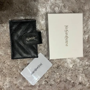 Svart plånbok från Yves Saint Laurent - Elegant svart plånbok i skinn med ett stilrent mönster och en YSL logga. Plånboken har en praktisk knappstängning i silver på framsidan. box och äktehetsbevis medföljer. Aldrig använd 💕💕