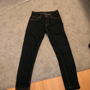 Mörkblåa jeans från Nudie Jeans - Snygga mörkblåa jeans från Nudie Jeans med orange sömmar. De har en klassisk femficksdesign och en knappgylf. Perfekta för en stilren look.