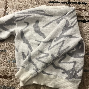 Vit och grå stickad tröja - Säljer en mysig vit och grå stickad tröja med abstrakt mönster. Tröjan har långa ärmar och ribbstickade muddar vid ärmslut och nederkant. Perfekt för kyliga dagar!