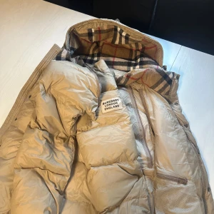 Beige dunjacka från Burberry - Snygg beige dunjacka från Burberry med klassiskt rutigt foder. Jackan har en dragkedja framtill och en hög krage för extra värme. Perfekt för kyliga dagar med stil.
