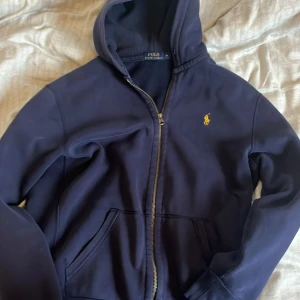 Mörkblå zip hoodie från Polo Ralph Lauren - Säljer en snygg mörkblå hoodie från Polo Ralph Lauren med dragkedja och det klassiska gula logotypen på bröstet. Perfekt för en avslappnad stil med sina långa ärmar och bekväma passform. Nypris 2000